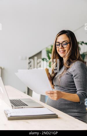 Junge, positive Frau erwartet ein Baby, das ein Kleid und eine Brille trägt und Heißgetränke trinkt und Papier liest, während sie von zu Hause aus mit einem Netbook arbeitet Stockfoto