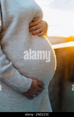 Gekürzte, nicht wiedererkennbare, schwangere Frau in warmem Kleid, die den Bauch berührt, wenn der Sonnenuntergang hell ist Stockfoto