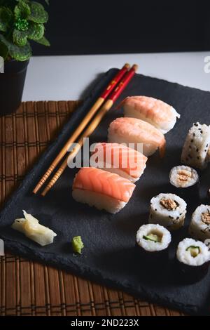 Leckere Sushi-Brötchen mit Fisch, serviert auf schwarzem Brett in der Nähe von Essstäbchen und Wasabi Stockfoto