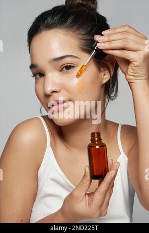 Pensive junge Frau mit weißem Oberteil, die Gesichtsöl aufträgt und dabei vor grauem Hintergrund steht Stockfoto