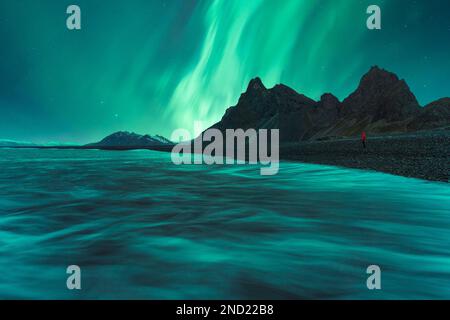 Rückblick auf anonyme Touristen, die an einer nassen Küste in der Nähe des ruhigen Meeres und rauer Berge stehen und den farbenfrohen Himmel mit Polarlichtern während des Ausflugs in Eystraho bewundern Stockfoto