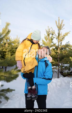 Glückliches Vorschulmädchen in warmer Kleidung und Hut, das während der Winterferien im verschneiten Wald auf der Schulter des Vaters sitzt Stockfoto