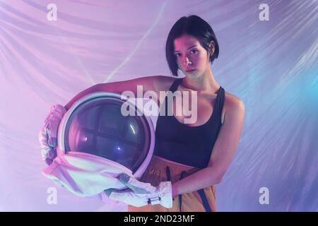 Eine ernsthafte junge Frau in legerer Kleidung, die in Neonlicht mit einem Kosmonautenhelm im Studio in die Kamera schaut Stockfoto