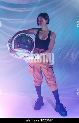 Eine seriöse junge Frau in legerer Kleidung, die im Neonlicht mit einem Kosmonautenhelm im Studio wegsieht Stockfoto