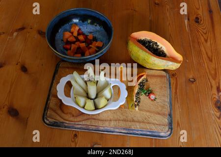 Zubereitung des Obstsalats – in Scheiben geschnittene Birne in einem Gericht mit Pepino und Erdbeeren, die hinzugefügt werden. Auf Holzbrett. Stockfoto
