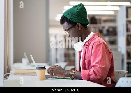 Fokussierter Schwarzer mit Brille, der in der Bibliothek oder im Klassenzimmer online auf einem Laptop lernt. Fernunterricht. Stockfoto