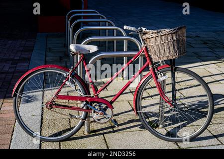 Ein gemütliches, altes, rotes Frauenrad ist in der Sonne an einen Fahrradständer gefesselt. Vorne ist ein Korb aus Korb, ein weißer Sitz und Handschlaufen. Stockfoto