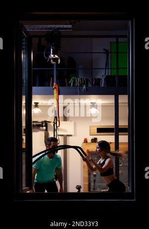 Fit Mädchen mit Training mit Seilen mit einem männlichen Fitness-Trainer in einem Fitness-Studio. Blick von außen. Stockfoto