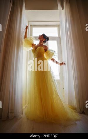 Die Silhouette einer Frau in einem goldenen, luxuriösen Kleid vor dem Hintergrund eines Fensters hält einen Vorhang mit den Händen. Elegante Dame in gelber Seide Stockfoto