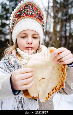 Russisches Nationalfestival Maslenitsa, Shrovetide. Kleine süße, wunderschöne Mädchen im Kopftuch essen große leckere Pfannkuchen, viel Spaß im Winter Pfannkuchen Urlaub Stockfoto