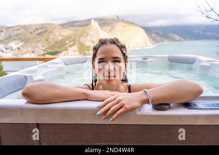 Nehmen Sie sich Zeit für sich. Badesachen im Freien mit Blick auf die Berge und das Meer. Eine Frau im schwarzen Badeanzug entspannt sich im Hotelpool und bewundert die Aussicht Stockfoto