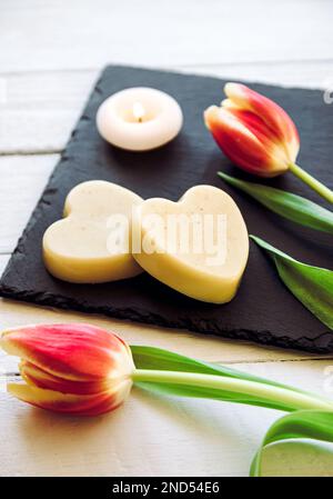 Selbstgemachte, feuchtigkeitsspendende Handcreme. Herzförmige Creme Bar aus allen natürlichen Zutaten. Schwarzer Steinboden mit frischen Tulpen. Stockfoto