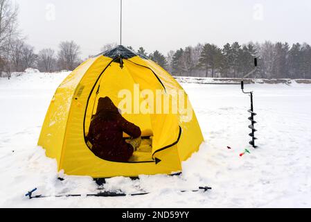 Anglermädchen winterten in der Kapuze und saßen in einem Zelt und Eisschrauben vor dem Hintergrund von Schnee und Wald in Russland. Stockfoto