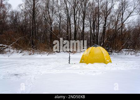 Gelbes Fischerzelt mit einem Eisloch im Winter auf dem Eis am Waldrand bei einem Schneesturm. Stockfoto