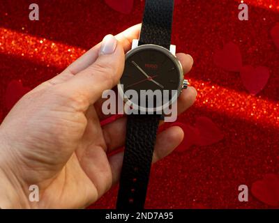 Hugo von Hugo Boss. Hand mit schwarzer Uhr vor Papierhintergrund mit rotem Herz. Grußkarte, präsent. Valentinstag-Urlaubskonzept. Flach aufgelegt w Stockfoto