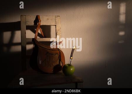 Eine Jägertasche aus Leder mit einer Vorderseite, dekoriert mit Hirschhaaren und einem Taschenmesser, in einen Apfel von Granny Smith gestochen, auf dem Sitz eines Sessels. Stockfoto