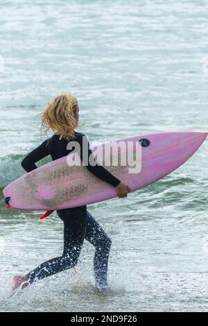 Eine eifrige junge Surferin, die ihr farbenfrohes Surfbrett im Fistral Beach in Newquay in Cornwall in England trägt. Stockfoto