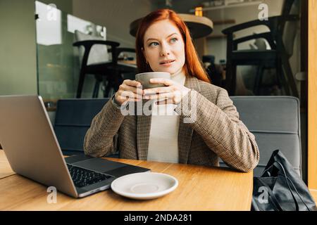 Eine Frau mit Ingwerhaaren, die eine Tasse hält und das Fenster in einem Café ansieht. Geschäftsfrau genießt ihren Kaffee. Stockfoto