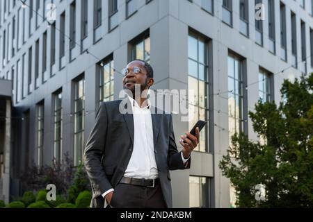 Foto von unten. Porträt eines afroamerikanischen Geschäftsmanns, der in einem Anzug vor einem Büro steht und ein Telefon in der Hand hält. Er hält seine Hand in der Tasche, sieht selbstbewusst zur Seite und lächelt. Stockfoto