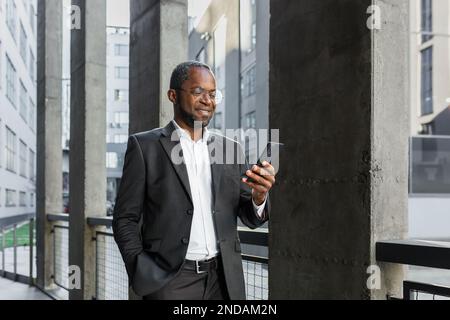 Porträt eines afroamerikanischen Geschäftsmanns, Banker, der in einem Anzug vor einem Bürogebäude steht und ein Telefon in der Hand hält. Er behält seine Hand in der Tasche, liest Nachrichten, schreibt eine Nachricht, checkt seine Post. Stockfoto