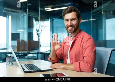 Porträt eines erfolgreichen jungen Programmierers, Designers, Freiberufler in einem roten Hemd, der im Büro an einem Laptop sitzt und arbeitet. Er sieht lächelnd in die Kamera. Zeigt mit der Hand OK an. Stockfoto