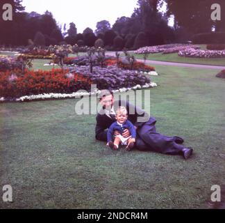 Vater und Sohn der 1950er. Der Vater trägt Anzug und Krawatte, der kleine Junge trägt Shorts und eine Strickjacke. Sie liegen auf Gras in einem formellen Park. Dieses Bild wurde aus der Originalfolie entnommen. Stockfoto
