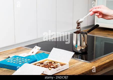 Zürich, Schweiz - Januar 7 2023: Lindt Mini Pralines Box. Offene kleine Geschenkbox mit Schweizer Schokoladensorten und eine Hand junger Frau, die türkischen Kaffee auf der Küchenplatte macht. Stockfoto