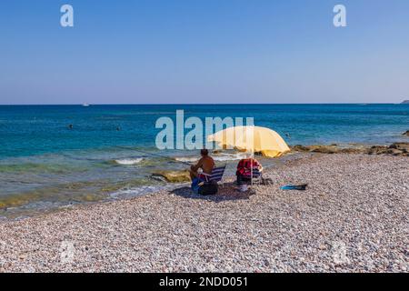 Wunderschöner Blick auf Männer beim Angeln an der Mittelmeerküste, die unter einem Sonnenschirm sitzen. Griechenland. Stockfoto