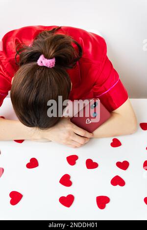 Das Mädchen ist traurig, legt ihren Kopf in die Hände, hält ein Smartphone mit Herz in einer Kirschtasche, wartet auf eine SMS oder eine Einladung zu einem Date. Stockfoto