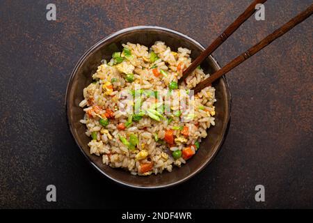 Authentischer chinesischer und asiatischer gebratener Reis mit Ei und Gemüse in einer braunen Keramikschüssel, Blick von oben, dunkler rustikaler Betontisch-Hintergrund. Traditionelles Gericht Stockfoto