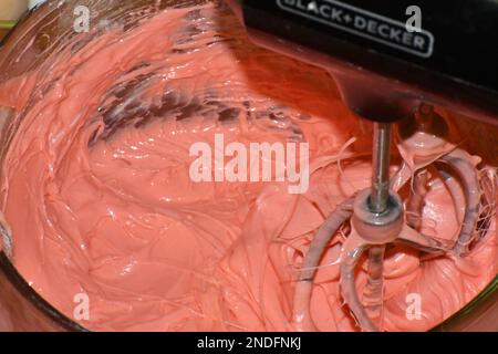 Ein Handmixer, der rosafarbenen Kuchenteig in einer Rührschüssel aus Glas in Bewegung mischt. Stockfoto