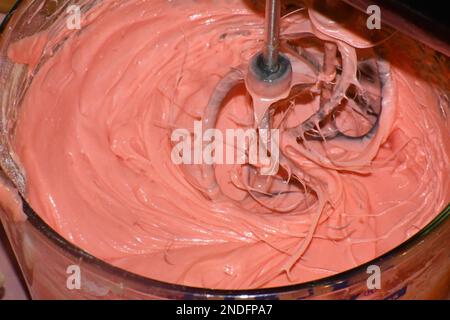 Ein Handmixer, der rosafarbenen Kuchenteig in einer Rührschüssel aus Glas in Bewegung mischt. Stockfoto