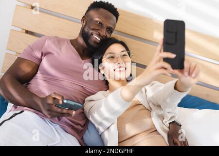 Glückliches, vielseitiges Paar, das im Bett liegt und Smartphones benutzt. Verbringen Sie Zeit zu Hause, in Ihrem Haushalt und in Ihrem Lebensstil. Stockfoto