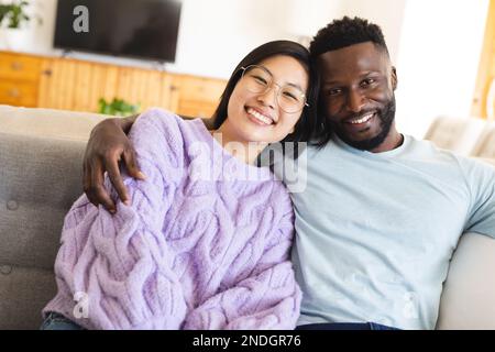 Portrait eines glücklichen, vielfältigen Paares, das auf dem Sofa sitzt und sich umarmt. Verbringen Sie Zeit zu Hause, in Ihrem Haushalt und in Ihrem Lebensstil. Stockfoto