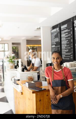Porträt einer fröhlichen, birassischen Barista, die eine Schürze im Café trägt. Café, Freizeit und Stadtkonzept. Stockfoto
