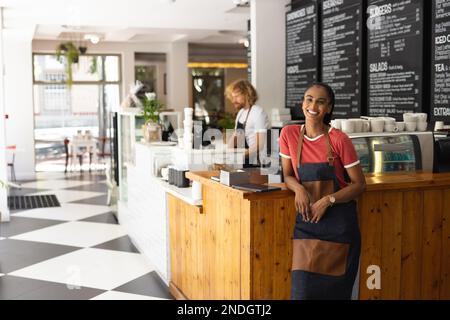 Porträt einer fröhlichen, birassischen Barista, die eine Schürze im Café trägt. Café, Freizeit und Stadtkonzept. Stockfoto