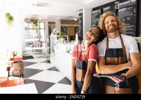 Porträt von glücklichen, vielfältigen Baristas, die Schürzen tragen und im Café ein Tablet verwenden. Café, Freizeit und Stadtkonzept. Stockfoto