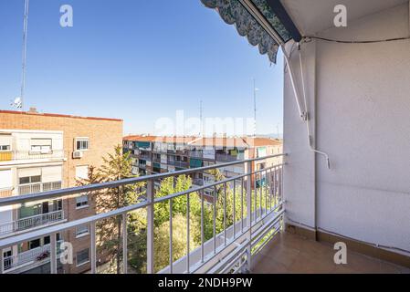 Terrasse eines Hauses mit weißem Metallgeländer und einer grünen, ausziehbaren Markise mit Mustern und Blick auf andere Gebäude in der Stadt Stockfoto