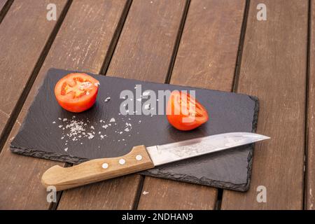 Zwei Teile einer halbreifen Tomate mit reifen Salzflocken auf einem glänzenden schwarzen Schieferbrett und einem Küchenmesser Stockfoto