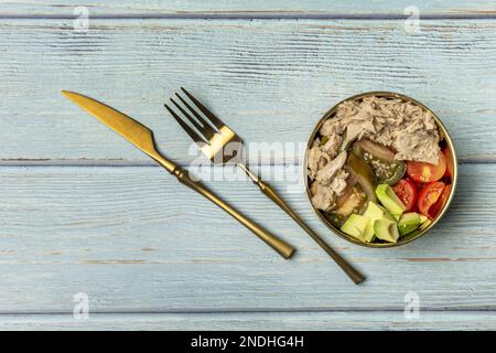 Ein wunderbarer Thunfischkonserven-Salat mit gewürfelten Tomaten und Avocados in einer goldenen Schüssel mit goldenem Besteck an der Seite auf einem blauen Tisch Stockfoto