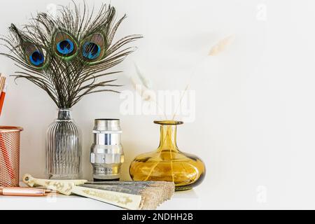 Dekorative Elemente auf einem weißen Regal mit Glasgefäßen, Vintage-Gläsern, Vasen mit Pfauenfedern und einem Ventilator Stockfoto