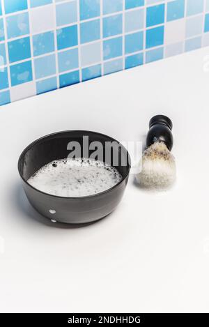 Ein schwarzer Behälter mit Schaumseife und eine Haarbürste zum Auftragen von Schaum auf das Gesicht, der auf einer weißen Oberfläche einer Toilette platziert wird Stockfoto