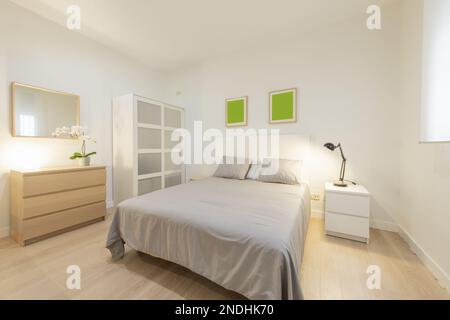 Doppelzimmer mit weißem Kopfteil aus Holz, grauen Bettüberwürfen und Kissen, weißem Nachttisch mit Klapplampe und EichenKommode mit passenden Schubladen Stockfoto