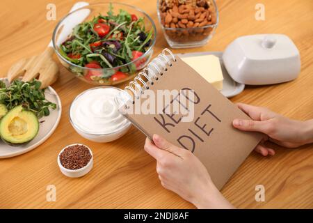 Frau mit Notizbuch mit Phrase Keto Diet und verschiedenen Produkten am Holztisch, Nahaufnahme Stockfoto