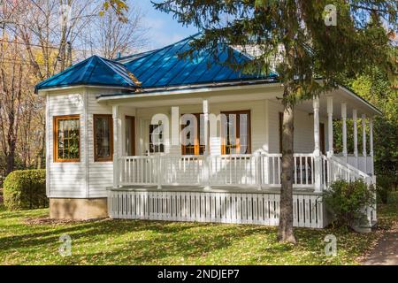 Alte weiße, horizontale Holzplanke verkleidete Hausfassade mit braunen Zierleisten und blauem Blechdach im Herbst. Stockfoto