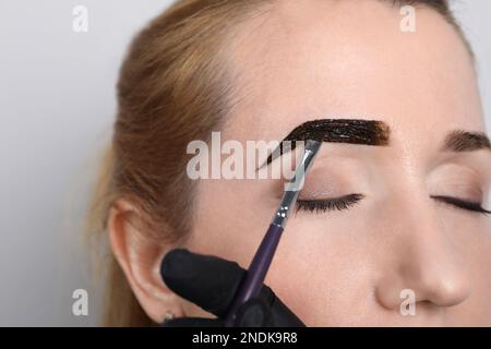 Kosmetikerin, die bei Augenbrauenkorrekturverfahren Farbtöne auf hellgrauem Hintergrund aufträgt, Nahaufnahme Stockfoto