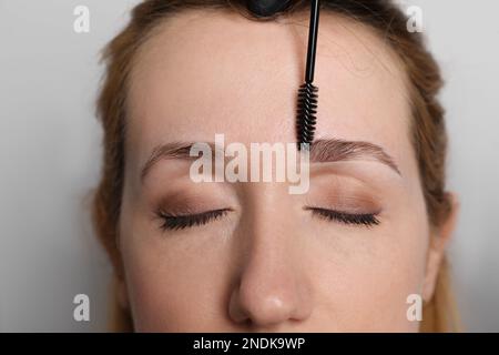 Kosmetikerin putzt die Augenbrauen der Frau, bevor sie auf hellgrauem Hintergrund abfärbt, Nahaufnahme Stockfoto
