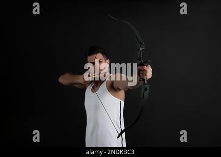 Mann mit Pfeil und Bogen, der Bogenschießen auf schwarzem Hintergrund praktiziert Stockfoto