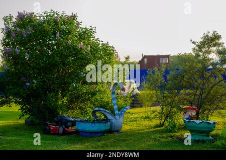 Die Gestalt einer hausgemachten Gans aus einem Autoreifen mit Blumenbeeten steht im Frühling auf einem Gartengelände unter Büschen auf grünem Gras. Stockfoto