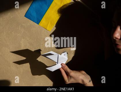 Papiertaube des Friedens in weiblicher Hand, Origami, Kontrastschatten und ukrainische Flagge. Konzept der Aufmerksamkeit auf den Krieg in der Ukraine. Spiel des Schattens und ich Stockfoto